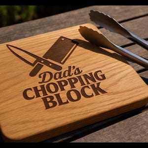Könnte beinhalten: Ein Holzschneidebrett mit der Gravur "Dad's CHOPPING BLOCK" sowie Messer- und Hackbeil-Illustrationen. Edelstahlzangen mit schwarzen Griffen liegen daneben. Das Brett ist aus hellbraunem Holz gefertigt.
