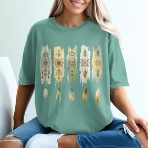 May include: A teal blue t-shirt with a graphic print of five watercolor-style vertical stripes with geometric patterns and feathers. The stripes are in shades of yellow, green, and brown.