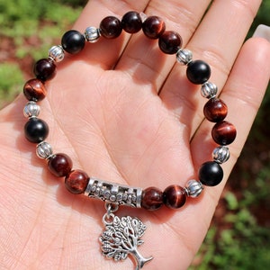May include: A beaded bracelet featuring alternating red-brown and black beads, interspersed with silver-colored spacers. A silver tree charm hangs from the bracelet. The bracelet is shown against a natural background.