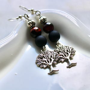 May include: Earrings with silver-tone tree charms, black and red beads, and silver-tone accents. The earrings are displayed on a white, glossy surface. The jewelry is designed for everyday wear.