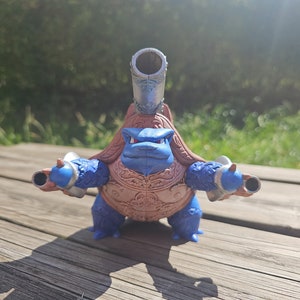 May include: A blue and brown toy turtle with cannons on its back and arms. The turtle is standing on a wooden surface.