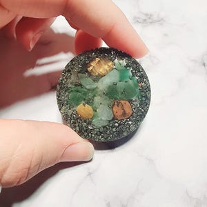 May include: A round, decorative object with a dark gray border. The center features green and tan stones, with a scattering of small, dark gray particles. The object is held by a hand against a white marble background.