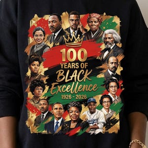 Könnte beinhalten: Schwarzes Sweatshirt mit einer farbenfrohen Grafik, die "100 Jahre Black Excellence" von 1926-2026 feiert. Das Design zeigt Porträts bemerkenswerter Persönlichkeiten vor einem Hintergrund in Rot, Schwarz und Grün.