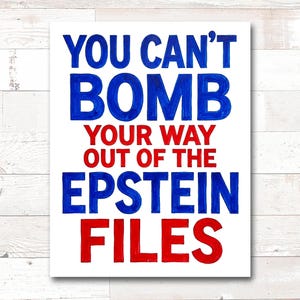 May include: A white rectangular sign with the text "YOU CAN'T BOMB YOUR WAY OUT OF THE EPSTEIN FILES" in blue and red lettering. The sign is against a white wooden background.