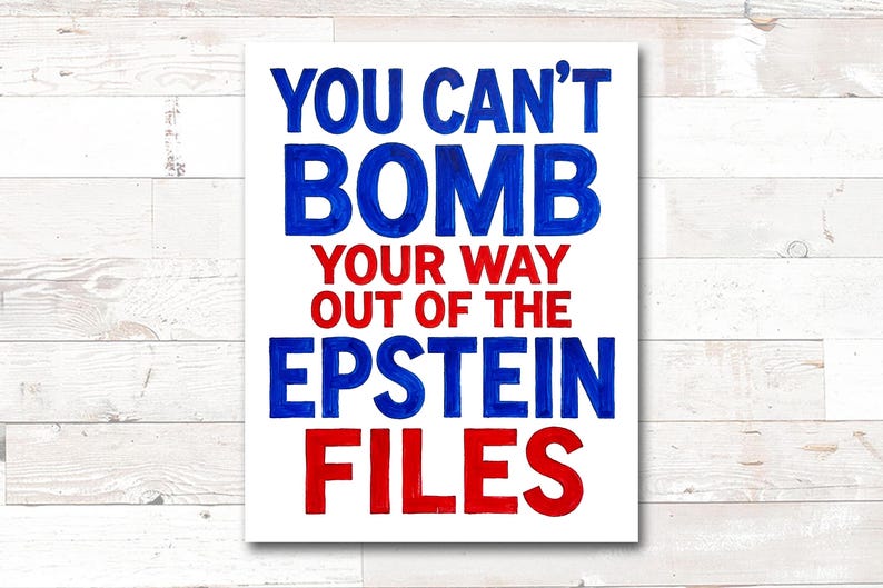 May include: A white rectangular sign with the text "YOU CAN'T BOMB YOUR WAY OUT OF THE EPSTEIN FILES" in blue and red lettering. The sign is against a white wooden background.