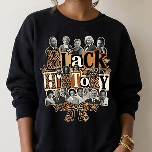 May include: Black sweatshirt featuring a graphic design with the words "Black History" in a mix of leopard print and solid colors. The design includes portraits of historical figures and a bow.