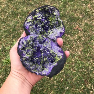 May include: Two purple and green geode halves with a sparkly, crystalline interior. The geode halves are held in a hand.