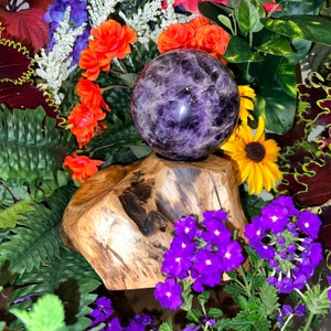 May include: A purple amethyst sphere resting on a wooden base surrounded by purple flowers and green foliage.