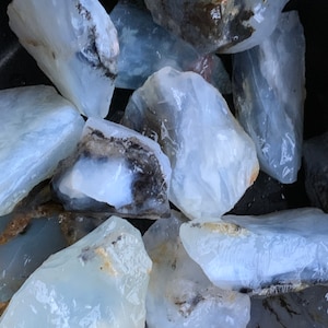 May include: A collection of white and gray opal rough stones. The stones are translucent and have a milky white color. Some of the stones have black markings.