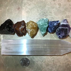 May include: A set of seven chakra stones, including black obsidian, red jasper, yellow citrine, green aventurine, blue sodalite, amethyst, and clear quartz, arranged in a line on a white selenite charging plate.