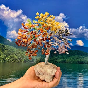 May include: A handcrafted tree sculpture with a silver wire trunk and branches, adorned with colorful gemstone leaves. The base is a rough-cut stone. The leaves transition from yellow to orange, red, purple, and blue. The background features a lake and sky.