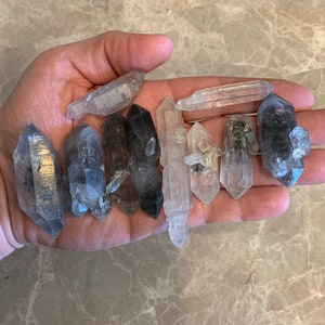 May include: A collection of ten clear quartz crystal points with some having black inclusions. The crystals are various sizes and shapes.