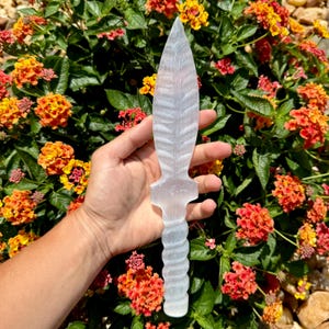 May include: A clear, carved selenite dagger with a pointed blade and a textured handle. The dagger is held in front of a backdrop of colorful orange and yellow flowers.