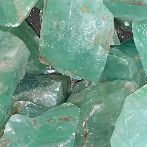May include: A close-up of a pile of green, translucent, rough-cut crystals. The crystals have a slightly cloudy appearance and some have brown inclusions.