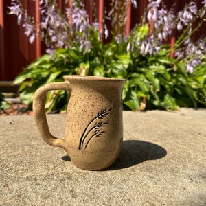 May include: A speckled tan ceramic mug with a black wheat design. The mug has a rounded handle.
