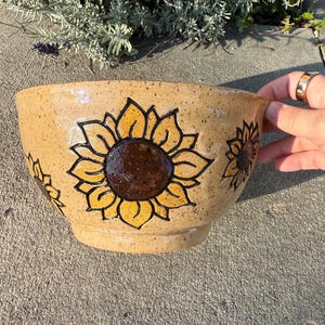 May include: A tan ceramic bowl with a sunflower design. The bowl features three sunflowers with yellow petals and dark brown centers. The bowl is round and has a wide opening. The background is a concrete surface.