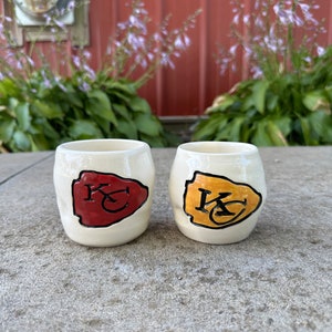 May include: Two white ceramic mugs with a red and yellow football-shaped design. The mugs have the letters "KC" in black on the design.