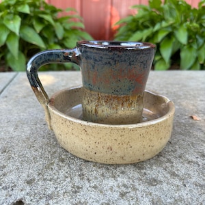 May include: A handmade ceramic mug with a matching saucer. The mug is dark blue and brown with a handle. The saucer is tan with dark speckles. The mug sits inside the saucer, creating a unique design.