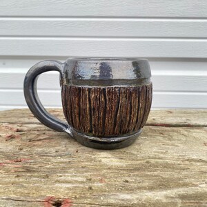 May include: A ceramic mug with a dark brown glaze and a wood grain design. The mug has a rounded body and a handle.