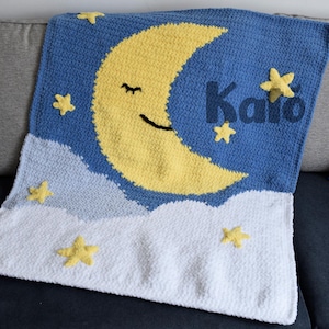 May include: A handmade blue, yellow, and white crochet blanket featuring a smiling yellow crescent moon, stars, and clouds. The name "Kato" is embroidered in blue. The blanket is folded on a gray couch.