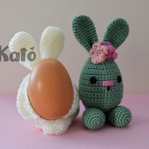 May include: Two crocheted Easter bunny ornaments. One is white and holds a brown egg. The other is green with pink flowers and a pink nose.