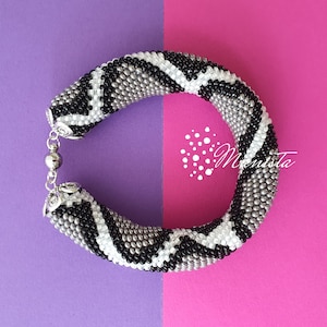 Puede incluir: Un brazalete de cuentas negras, blancas y plateadas con un patrón de piel de serpiente. El brazalete está sobre un fondo morado y rosa.