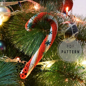 Puede incluir: Un adorno navideño de bastón de caramelo con cuentas rojas, blancas y verdes. El adorno cuelga de un árbol de Navidad decorado con luces y adornos. El texto "PDF PATTERN" es visible.