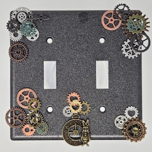 May include: A decorative, steampunk-style double light switch cover. The dark gray cover is embellished with various metal gears in shades of copper, bronze, silver, and green. A clock charm with a rabbit figure adds a whimsical touch to the design.