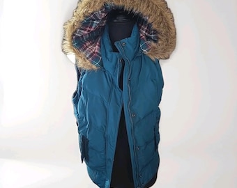 May include: A teal puffer vest featuring a hood with a brown faux fur trim. The interior of the hood is lined with a red, green, and white plaid fabric. The vest has a front zipper closure with a snap button placket and two side pockets.