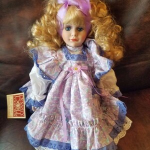 May include: A porcelain doll with blonde curly hair wearing a purple and white floral dress with a lavender ribbon and a purple bow in her hair. The doll is sitting on a brown leather surface. The doll is part of the Dynasty Doll Collection.