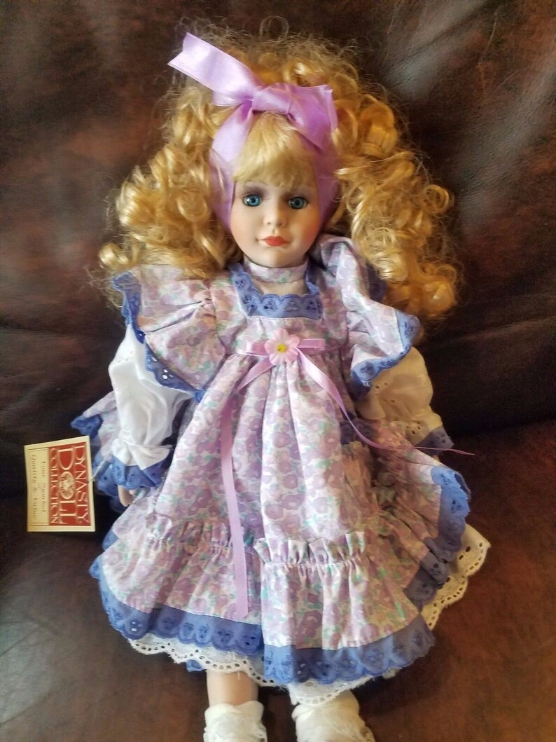 May include: A porcelain doll with blonde curly hair wearing a purple and white floral dress with a lavender ribbon and a purple bow in her hair. The doll is sitting on a brown leather surface. The doll is part of the Dynasty Doll Collection.