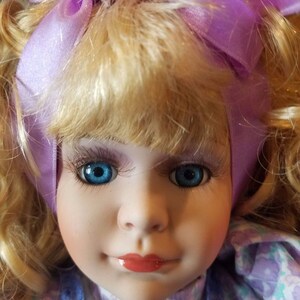 May include: A porcelain doll with blonde hair and blue eyes. The doll is wearing a purple ribbon and a floral dress.