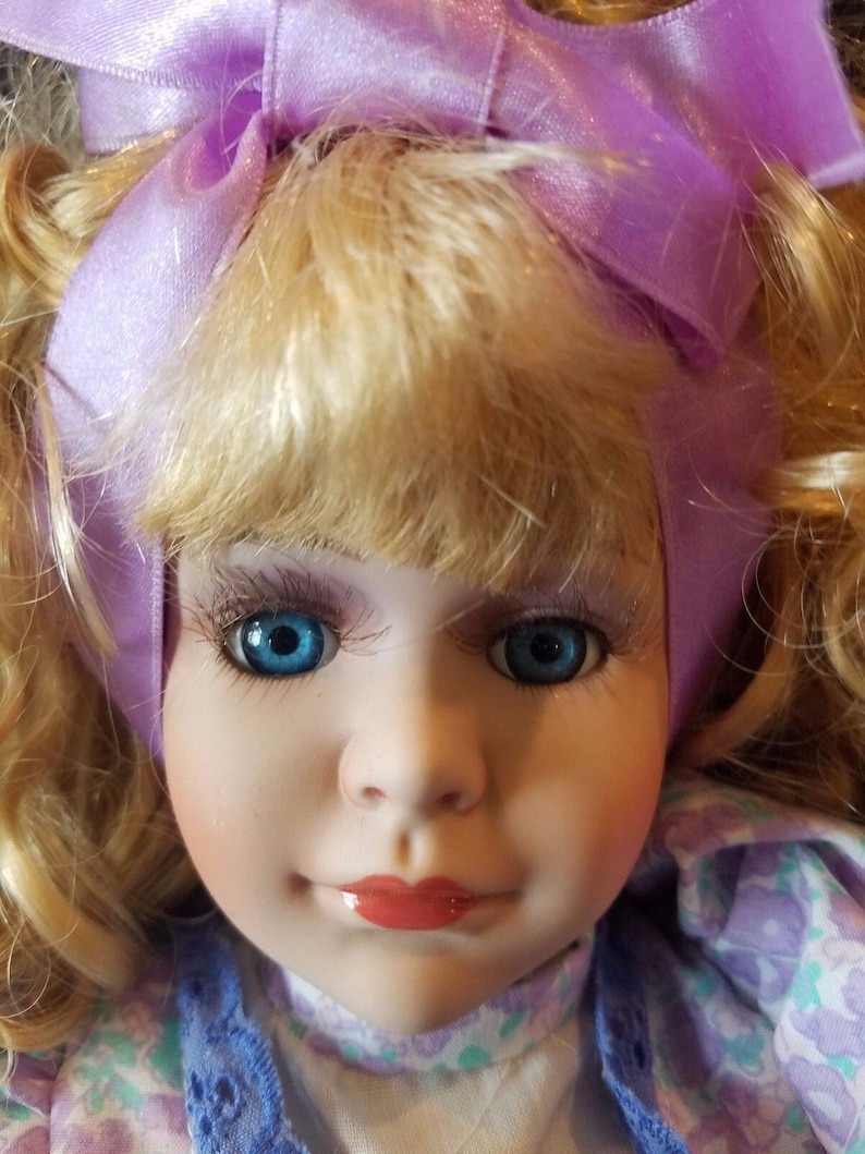 May include: A porcelain doll with blonde hair and blue eyes. The doll is wearing a purple ribbon and a floral dress.