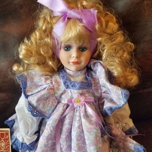 May include: A porcelain doll with blonde curly hair wearing a purple and white floral dress with a purple ribbon and a flower accent. The doll is wearing a purple bow in her hair. The doll is labeled "Dynasty Doll".