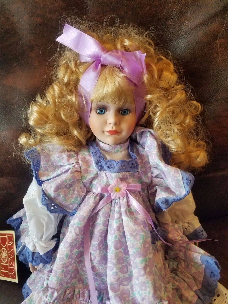 May include: A porcelain doll with blonde curly hair wearing a purple and white floral dress with a purple ribbon and a flower accent. The doll is wearing a purple bow in her hair. The doll is labeled "Dynasty Doll".