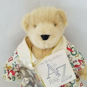 May include: A plush teddy bear with a light brown fur. The bear wears a white collared shirt with a colorful alphabet print and blue shoes. A tag reads "The Reading Collection A to Z by J. Vide B." and includes a pair of small glasses.