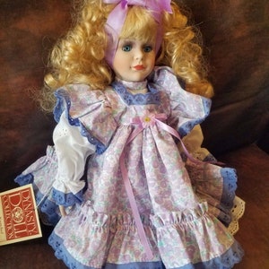 May include: A porcelain doll with blonde curly hair wearing a purple and white floral dress with a lavender ribbon and a purple bow. The doll is sitting on a brown leather surface. The doll is labeled "Dynasty Doll Collection" on a tag.