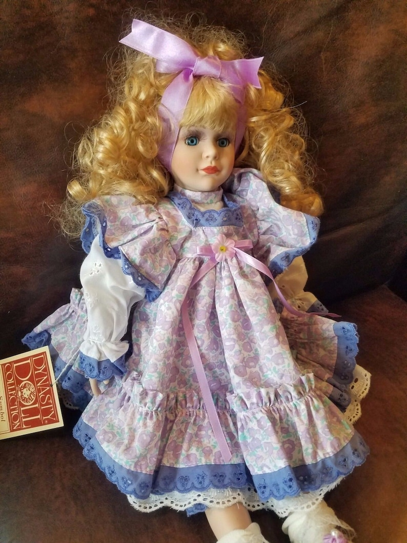 May include: A porcelain doll with blonde curly hair wearing a purple and white floral dress with a lavender ribbon and a purple bow. The doll is sitting on a brown leather surface. The doll is labeled "Dynasty Doll Collection" on a tag.