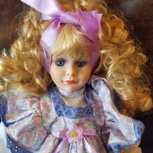 May include: A porcelain doll with blonde curly hair wearing a purple and white floral dress with a lavender ribbon and a matching bow in her hair.