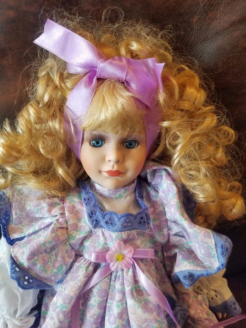 May include: A porcelain doll with blonde curly hair wearing a purple and white floral dress with a lavender ribbon and a matching bow in her hair.