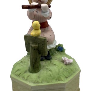 Puede incluir: Figura de porcelana de una niña tocando la flauta. Viste un vestido rosa con cinturón rojo y zapatos negros. Un pollito amarillo está sentado en un letrero. La figura está sobre una base verde con borde de madera.