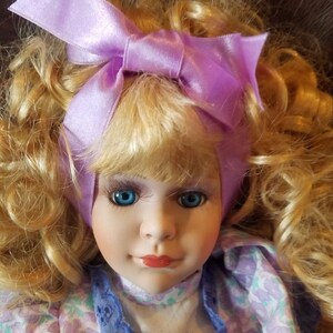 May include: A porcelain doll with blonde curly hair wearing a purple satin bow and a lavender and white floral dress with blue lace trim.