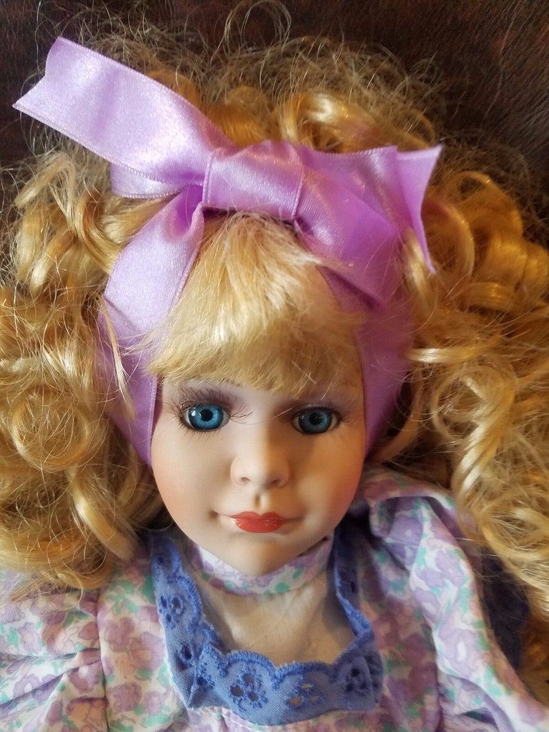 May include: A porcelain doll with blonde curly hair wearing a purple satin bow and a lavender and white floral dress with blue lace trim.