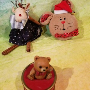 May include: A festive assortment of Christmas decorations. Includes a reindeer figure with cinnamon stick legs, a bunny wearing a Santa hat, and a small teddy bear on a red decorative box. The reindeer has a blue dress with white stars.