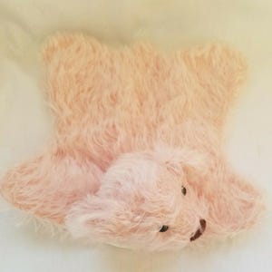 May include: A light pink, fluffy rug shaped like a teddy bear. The rug has a soft, shaggy texture and features a bear head with small black eyes and a brown nose. The bear's ears and paws are also visible.