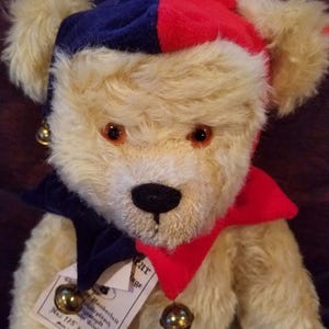 May include: A vintage teddy bear with a cream-colored plush body, wearing a jester's hat in red and blue, and a red collar. The bear has a black nose, brown eyes, and a tag with text and a bell.