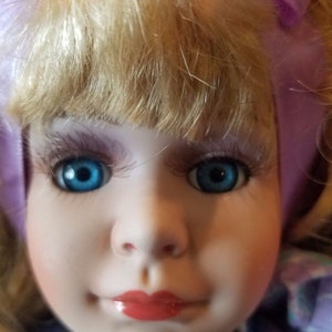 May include: A porcelain doll with blonde hair, blue eyes, and a purple bow. The doll is wearing a blue and purple dress with a floral pattern.