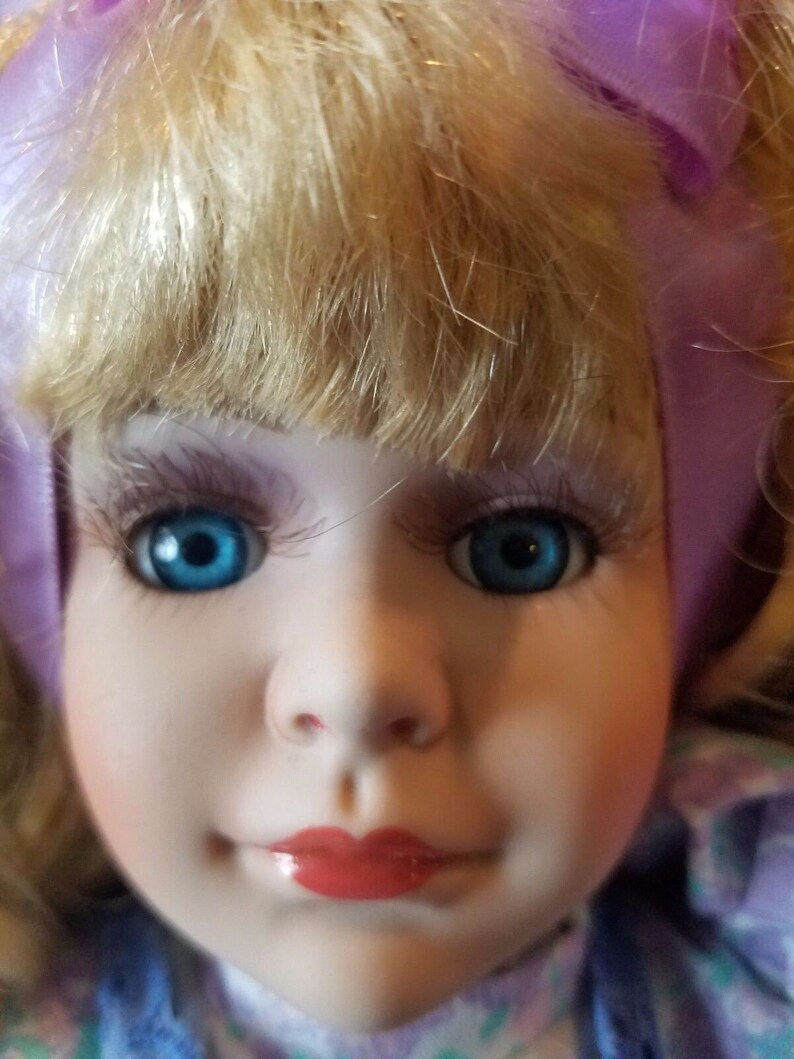 May include: A porcelain doll with blonde hair, blue eyes, and a purple bow. The doll is wearing a blue and purple dress with a floral pattern.