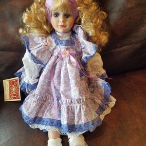 May include: A porcelain doll with blonde curly hair wearing a purple and white floral dress with a lavender ribbon and a matching headband. The doll is wearing white shoes with purple flowers.