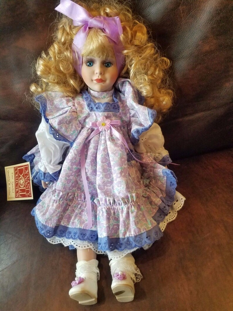 May include: A porcelain doll with blonde curly hair wearing a purple and white floral dress with a lavender ribbon and a matching headband. The doll is wearing white shoes with purple flowers.
